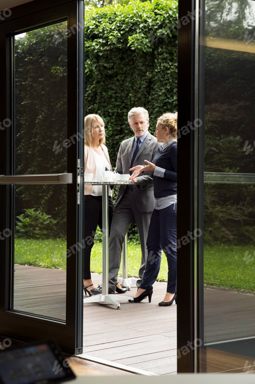 Preview: View through doorway of colleagues standing at patio table enjoying refreshments