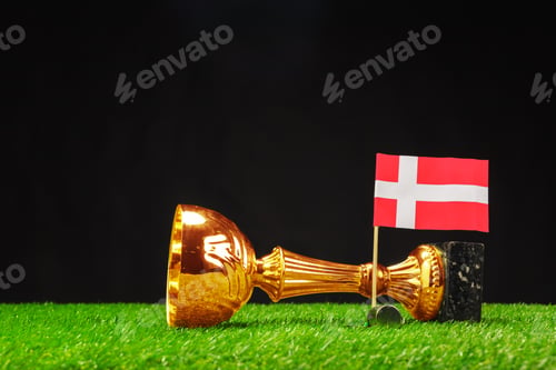 Preview: Golden trophy lies on green turf beside small flag representing Denmark in a celebratory scene