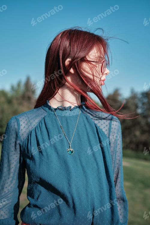 Preview: Teenage girl with red hair wearing glasses and blue blouse standing outdoors on a sunny day