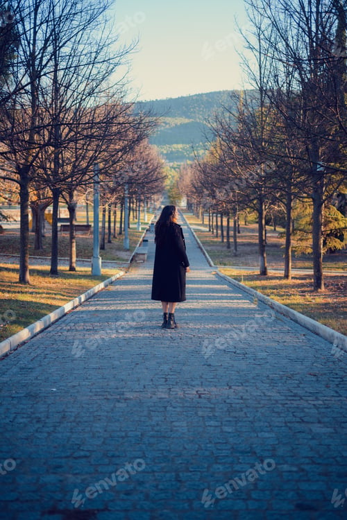 Preview: Woman in an overcoat and scarf walks through an autumn park at dusk