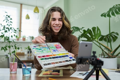Preview: Teenage guy drawing with watercolors, watching video class on smartphone