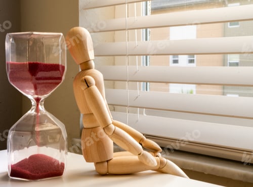 Preview: Closeup shot of an hourglass and a wooden human figure sitting by the window and waiting