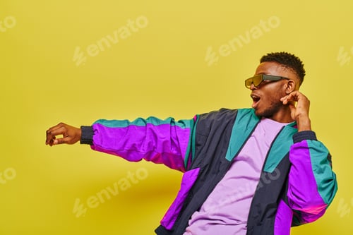 Preview: fashionable and energetic african american man in bright jacket and sunglasses dancing on yellow