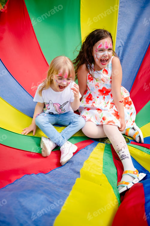 Preview: Two excited laughing girls having fun in multicoloured play parachute on kid’s birthday party.