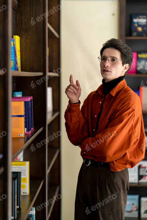 Preview: young guy in glasses stands near the bookshelves thoughtfully chooses a book in university library