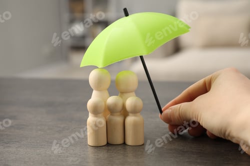 Preview: Woman holding umbrella over human figures at grey table indoors, closeup. Insurance concept