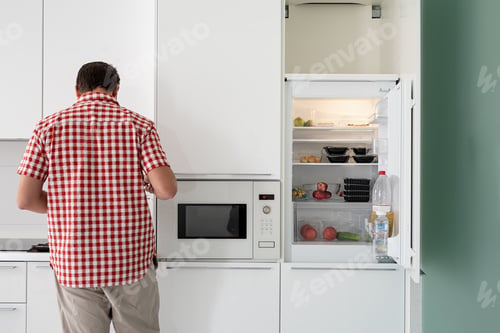 Preview: Young man makind tea standing next to the open fridge.
