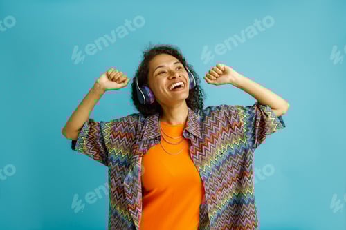 Preview: Smiling woman is enjoy music in headphones with raised hands standing on blue studio background