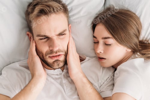 Preview: top view of exhausted man plugging ears with hands while lying in bed near snoring wife