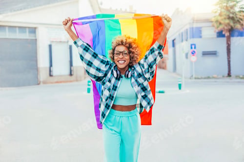 Preview: Black afro latin american woman holds up rainbow flag of gay pride outdoors