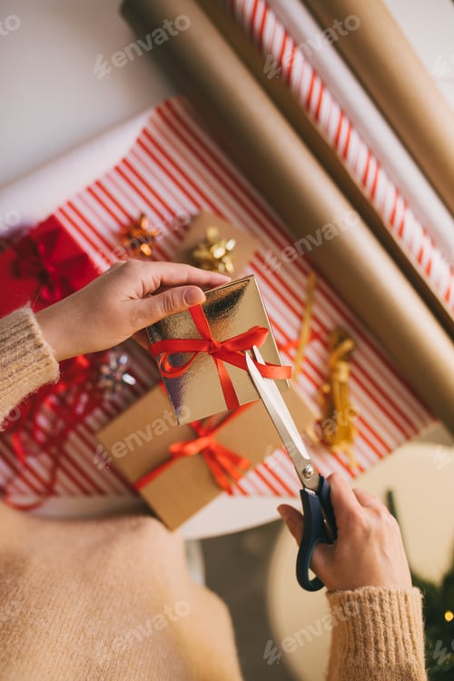 Preview: Young woman in sweater wrapping Christmas presents using paper, scissors and colorful ribbons.