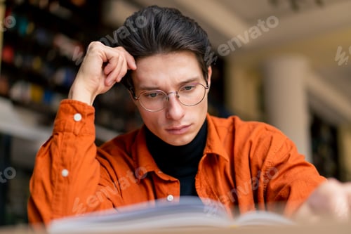 Preview: a young guy with glasses is sitting in library cafe and carefully reading a book about hobbies