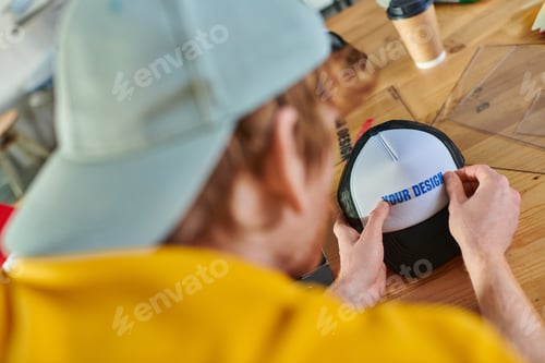 Preview: Blurred young designer holding printing layer near snapback while working near blurred coffee to go