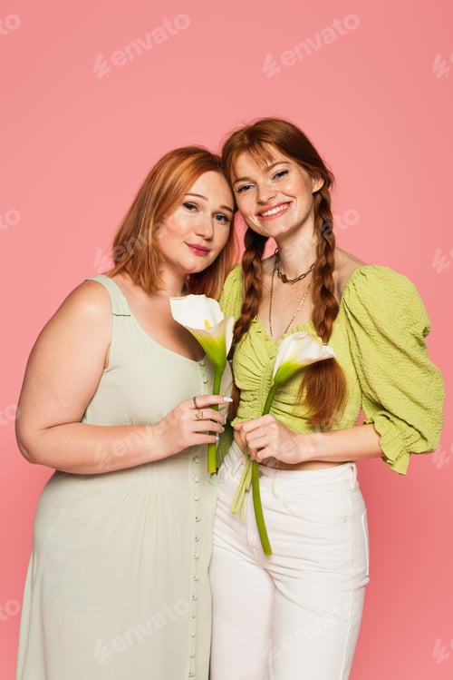 Preview: Smiling freckled woman holding calla lily near plus size friend isolated on pink