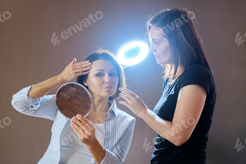Preview: Talking and smiling women beautician and mature female patient with mirror in beauty salon