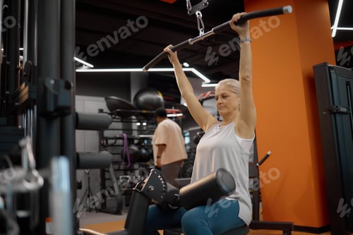 Preview: Senior Caucasian Woman Exercising on Lat Pulldown Machine in Modern Gym