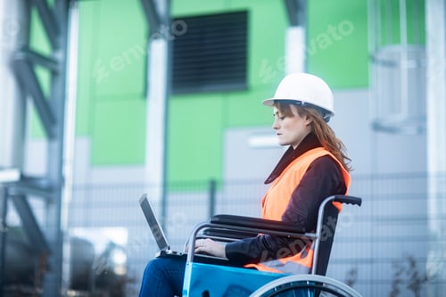 Preview: Young technician with safety helmet and vest in wheelchair working on laptop outdoors