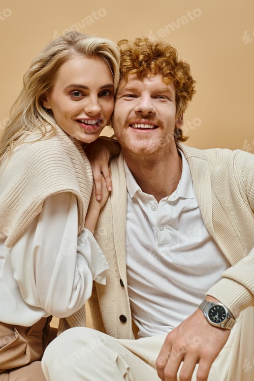 Preview: pleased blonde woman and redhead man in light-colored outfit looking at camera on beige backdrop