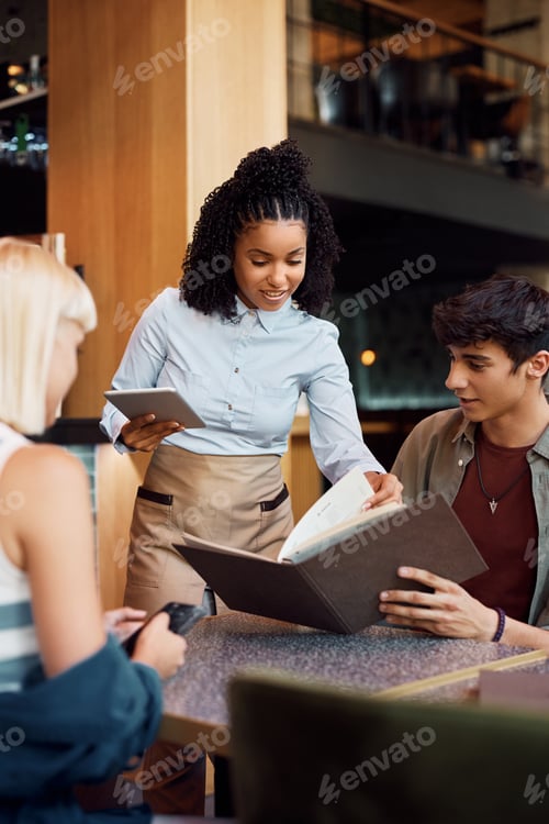 Preview: Happy black waitress showing a menu to guests in restaurant.