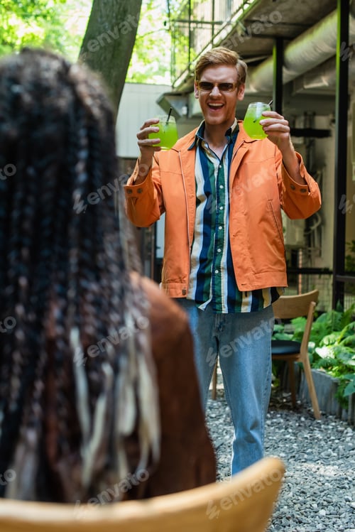 Preview: cheerful redhead man in sunglasses holding glasses with green alcohol cocktail near blurred friend