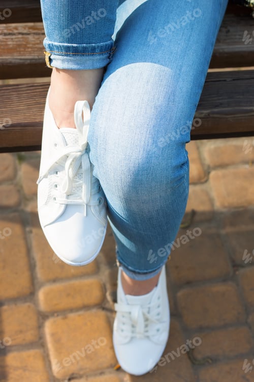 Preview: White sneakers on female legs in blue jeans on the asphalt background.