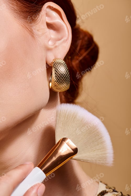 Preview: cropped redhead woman in gold earring applying bronzer with makeup brush on beige background