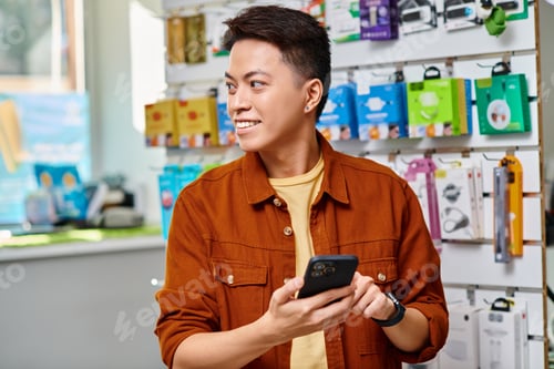 Preview: pleased asian entrepreneur holding mobile phone and looking away in private electronics shop