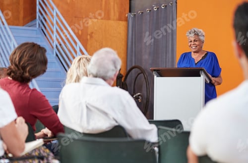 Preview: Senior Woman At Podium Chairing Neighborhood Meeting In Community Centre