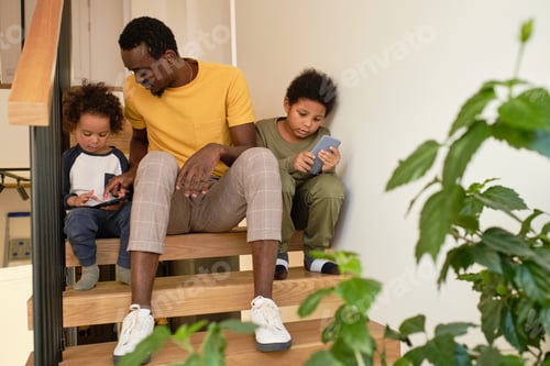 Preview: Young man of African ethnicity talking to one of his cute sons with smartphones