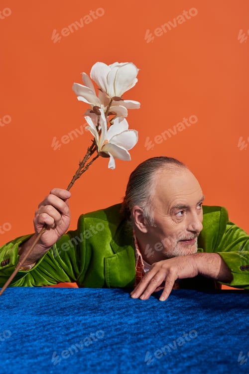 Preview: joyful and grey haired senior man sitting at table with blue velour cloth, holding white orchid
