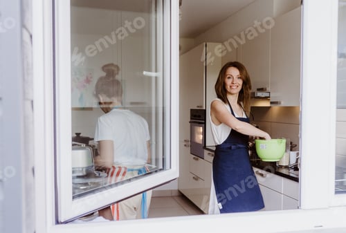 Preview: View from outside through open window at mother and teen daughter cooking at home together