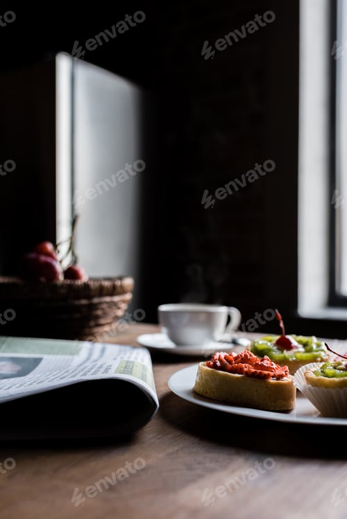 Preview: Still life of newspaper, breakfast with cakes and hot coffee on kitchen table in front of window
