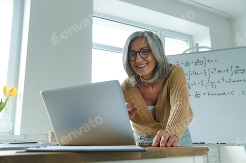 Preview: Mature woman teaching mathematics while standing near the whiteboard and looking at laptop
