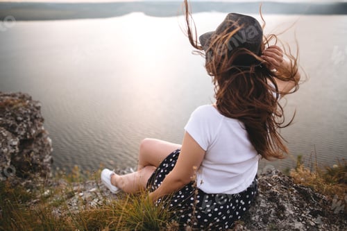 Preview: Traveler hipster girl in hat and windy hair sitting on top of rock mountain
