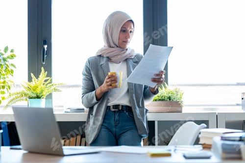 Preview: Beauty muslim business woman wearing hijab using her smart phone while holding papers in the office.