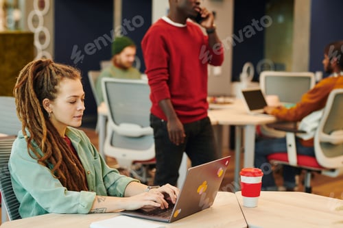Preview: Young Caucasian businesswoman with dreadlocks sitting by desk in front of laptop