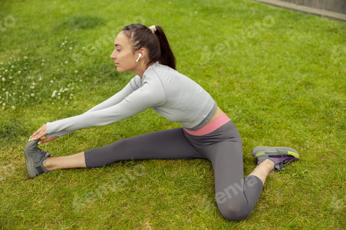 Preview: Beautiful young woman doing stretching exercise on green grass at city park