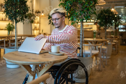 Preview: Calm disabled man in coffee shop sitting in wheelchair at table opening laptop getting ready to work