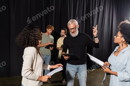 Preview: bearded art director pointing with finger while reading screenplay near multiethnic actresses