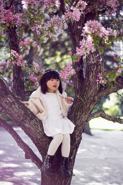 Preview: Korean girl in a white light fur coat and a headband sitting on a tree branch in a garden