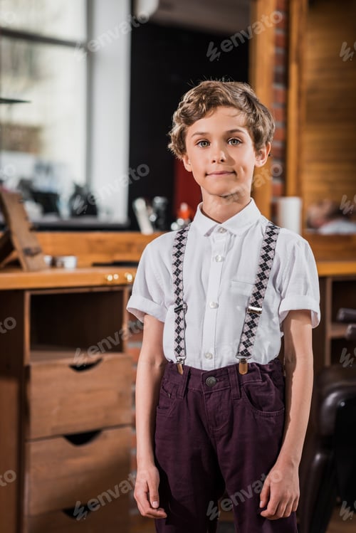 Preview: stylish little kid in white shirt with suspenders looking at camera at barbershop