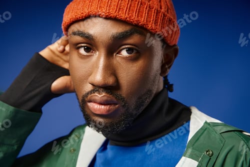 Preview: portrait of handsome african american man in knitted hat and winter attire posing on blue backdrop
