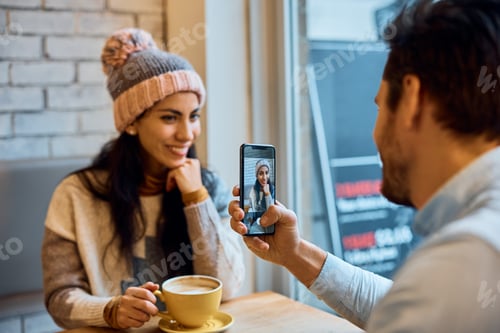 Preview: Close up of man photographing his girlfriend with smart phone in a cafe.