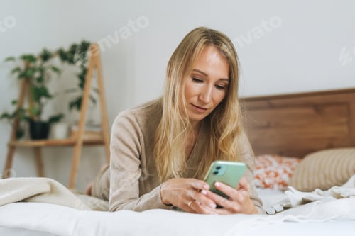 Preview: Beautiful blonde young woman using mobile on bed