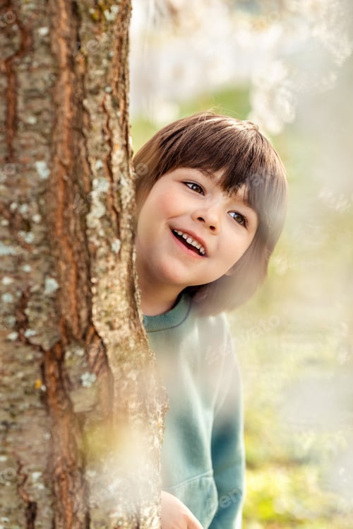 Preview: Little happy toddler boy playing hide and seek behind the tree in spring park.