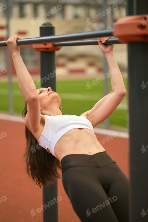 Preview: A fit woman performs a chin-up on an outdoor gym bar, her muscles straining with effort. This image