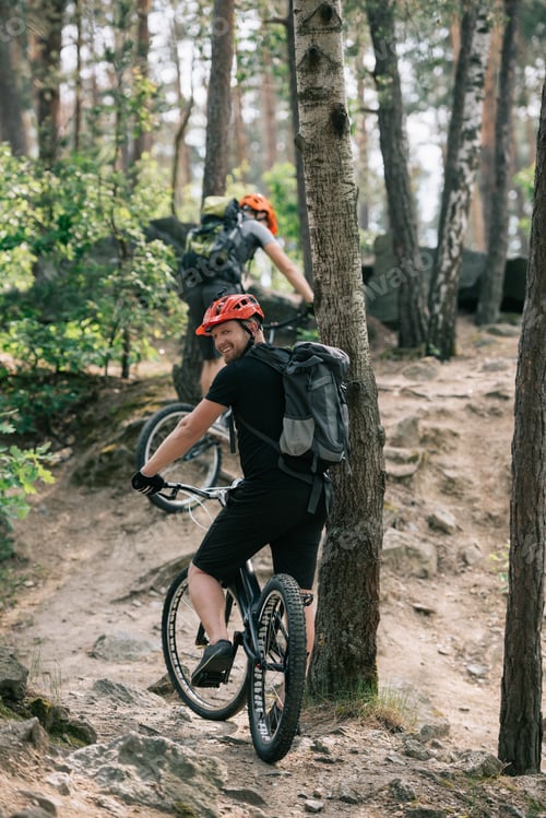 Preview: smiling male extreme cyclist riding on mountain bicycle with friend in forest