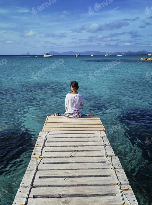 Preview: Vertical shot of a female sitting on a dock and admiring the turquoise seascape