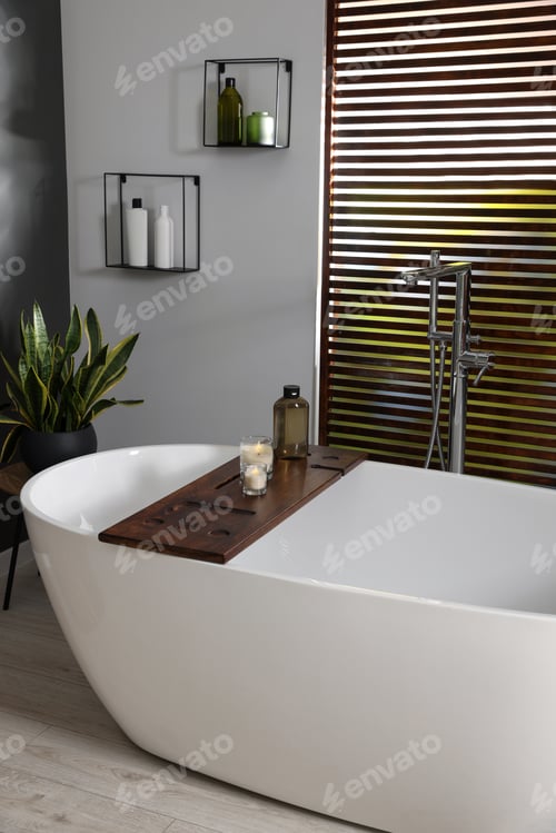 Preview: Stylish bathroom interior with ceramic tub, candles and care products on wooden bath tray