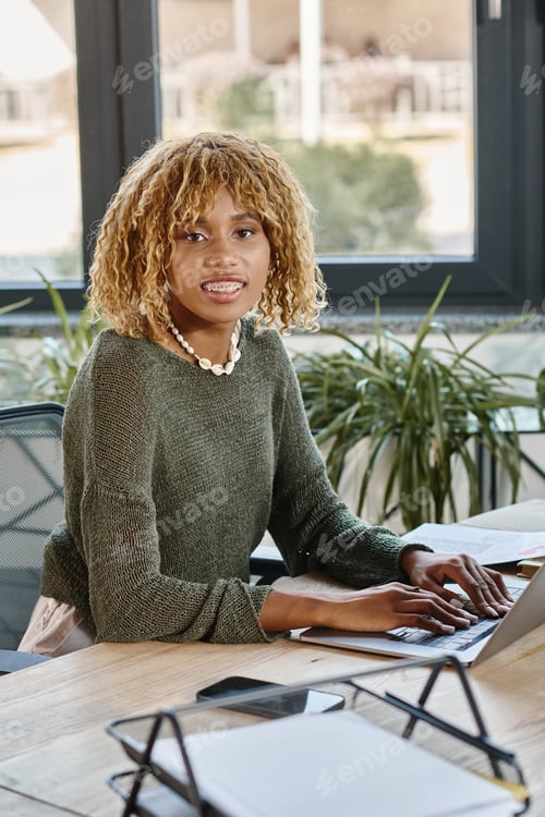 Preview: portrait of curly haired woman with braces smiling at camera with laptop on table, working process